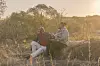 Chris and Dumi sit on a rock in a sunlit field in Botswana, sharing a quiet moment in a scene from Matabeleland, directed by Nyasha Kadandara.