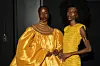 Two Black female models dressed in yellow, Afro-centric clothing, stand together backstage. The model on the left has short hair while the model on the left has a big afr