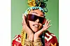 Doowap has colorful braided hair, oversized sunglasses, and stacked gold bangles, and she poses with her hands framing her face against a green background.