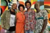 Three men and one woman dressed in colorful clothes smile directly into the camera, standing in front of a wall with colorful prints.