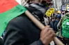 A protest scene in a city, with a blurred figure in the foreground holding a red, black, and green flag. In the background, demonstrators hold signs demanding immigrant rights and calling for the release of detained individuals, with buildings towering over the crowd.