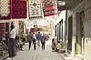 A bustling street market in Algeria in 1969, with people walking along a narrow cobblestone alley lined with shops displaying colorful woven rugs and carpets hanging overhead.
