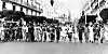 Black-and-white photo of a group of women in traditional African and Western attire walking arm-in-arm down a street in Algeria, with crowds watching from nearby buildings.