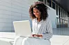 A portrait of a smiling young businesswoman using a laptop while sitting outside an office building.