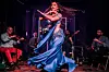 A bellydancer wearing a turquoise belly-dancing outfit is performing with long hair flying in front of a traditional ensemble of four musicians, playing the violin, tabla, saxophon,e and guitar.