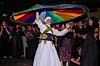 A traditional Arab dancer is wearing a white semazen dress with red embroidery and a black belt with golden embroidery, twirling a rainbow-colored textile above his head.