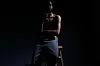 Tim Lyre sits on a bar stool, shirtless and wearing jeans, with a very dark background.