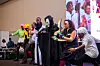 A diverse group of cosplayers in various costumes, including a character with a scythe, a character with twin blades, and others in colorful outfits, strike poses on stage at the cosplay competition during NAICCON.