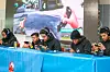 A group of gamers wearing headphones concentrate on their mobile devices during a gaming competition at NAICCON, with the game display on a large screen in the background.