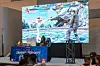 A gamer, wearing headphones and seated at a gaming desk, competes in the Swahili Esports Championships at NAICCON. A large screen behind her displays the intense in-game action from a fighting game, with spectators watching nearby.