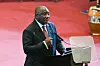 An older man, Cyril Ramaphosa, in a suit stands at a podium while speaking into a microphone. He stands against a red-carpeted background.