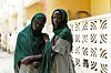 Twelve-year-old twin girls wearing green hijabs and black and white striped dresses, looking directly into the camera. One places her hand on the other\u2019s shoulder.