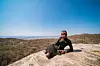 A Hadza girl is laying on a rock plateau, holding a camera and looking critically at the camera. Behind her, the vast landscape of Tanzania’s nature.