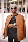 Wizkid wearing a black t-shirt and pants, a brown jacket, a gold chain with a Chanel logo pendant, a gray beret, and sunglasses.