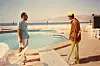 Two men stand by a pool, one wearing leisurewear and the other work clothes. Behind them, the Red Sea and the Red Sea Mountains.