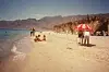 Several people are enjoying themselves on the beach. There's a red Coca-Cola umbrella and some straw huts; the Red Sea Mountains are in the background.