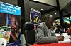 Ngũgĩ wa Thiong'o sits at a table signing a book during a public event, with a banner behind him displaying “Weep Not, Child Book Signing by Prof. Ngũgĩ wa Thiong’o” and a crowd of people gathered in the background.