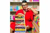 A smiling woman in a red dress and glasses seated beside a tall stack of books.