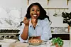Remi Idowu smiling while eating pasta in a modern kitchen, wearing a blue striped shirt with fresh herbs and cooking ingredients visible on marble countertop.