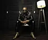 Nigerian star Olamide sits in a dark room dressed in all black leather and snake skin cowboy boots.