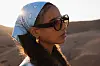 Profile shot of Rania Malek standing in the desert, wearing brown sunglasses, golden earrings and a blue bandana that ties back her long hair.