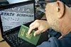 A man wearing glasses and a navy shirt holds his passport in front of his computer, displaying a Schengen visa