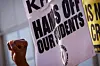 A close-up of a protest sign that reads “HANDS OFF OUR STUDENTS” in bold black letters, held beside a raised fist, symbolizing resistance and solidarity.