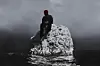 A lone figure wearing dark clothes and a red mask sits atop a white rock surrounded by dark, choppy waters, under a gloomy, overcast sky that evokes a sense of isolation and mystery.