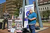 An older woman speaks into a microphone during a public event in Johannesburg, South Africa, advocating for library access. She stands beside a banner that reads \u201cLibraries are essential services,\u201d while holding a petition. Behind her, another man stands near posters titled \u201cPetition in a Book,\u201d filled with portraits of supporters.