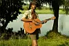 Tommy W\u00c1, photographed in soft natural light, holds a light brown-coloured guitar. He is wearing shorts and an unbuttoned shirt, and is standing in front of trees that barricade a lake.
