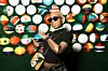 A woman poses in front of a vibrant backdrop made of balls decorated with African national flags. She's wearing a black top with black sunglasses and has a bold 'ROCKSTAR' tattoo on her arm.