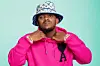 Kabza De Small wears a pink AMI Paris Ami de Coeur wool cardigan. He is also wearing an off-white and blue LV bucket hat, and is holding the collar of his sweater.