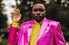 Willis Chimano stands confidently in a striking metallic pink suit with flared trousers and bold eye makeup.