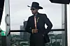 Ugo Mozie poses on a balcony wearing a black suit and hat.