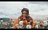 A still of Tamy Moyo in the visualizer for her latest single. She is in a garden of roses, gazing straight at the camera while wearing an orange dress.