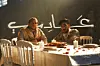 Two men sit at a table with a white tablecloth and red flowers, and white candles. Behind them, the word 3ady is written in Arabic in front of a black wall.