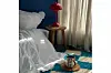 A white bed on the floor on top of a blue and white carpet. Next to it are a red lamp and a ceramic mug full of coffee.