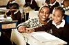 A teacher and her girl student are smiling into the camera in a classroom where the other students are focused on their work and writing.