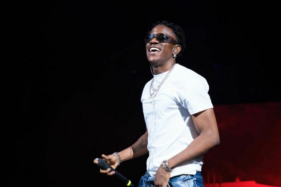 Victony performing live on stage with a mic. He wears a white t-shirt and blue jeans.
