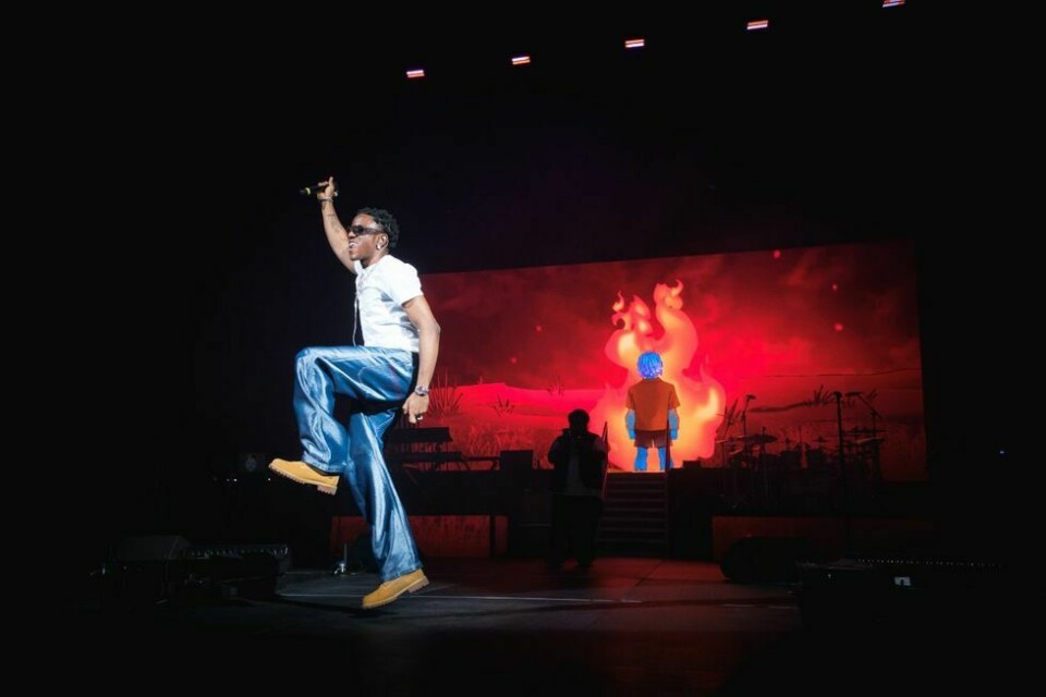 Victony performing live on stage with a mic. He wears a white t-shirt and blue jeans.